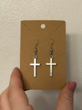 Simple silver cross earrings with hooks on brown paper earring holder, held against a white wall