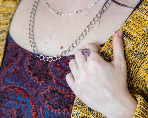 Model wearing chunky Amethyst claw style ring on index finger. She has colorful apparel, and is silver chains on neck.