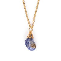 Rough cut sodalite with glue on bail, hanging on gold chain. Image has solid white background.
