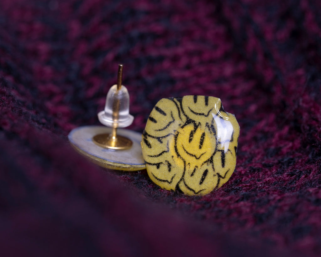 Creative image of smiley earrings, showing post sticking upright, and melting smiley earring facing forward, on burgundy and black fabric. 