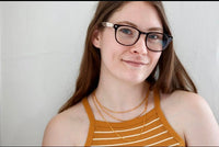 Female model smiling, wearing 3 layer gold chain with mustard yellow tank and glasses.